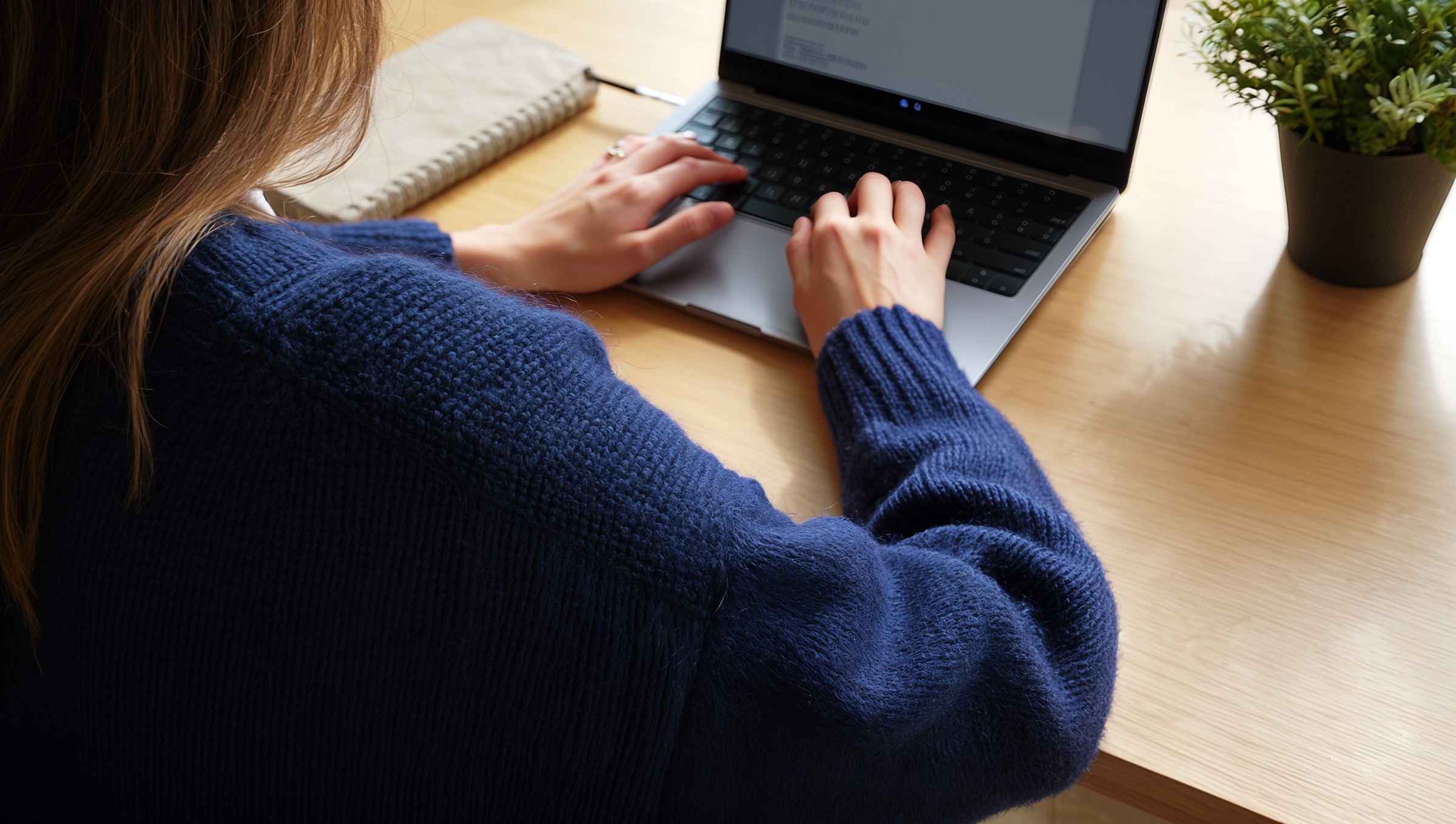 Une personne en pull bleu tape sur un ordinateur portable à un bureau en bois.